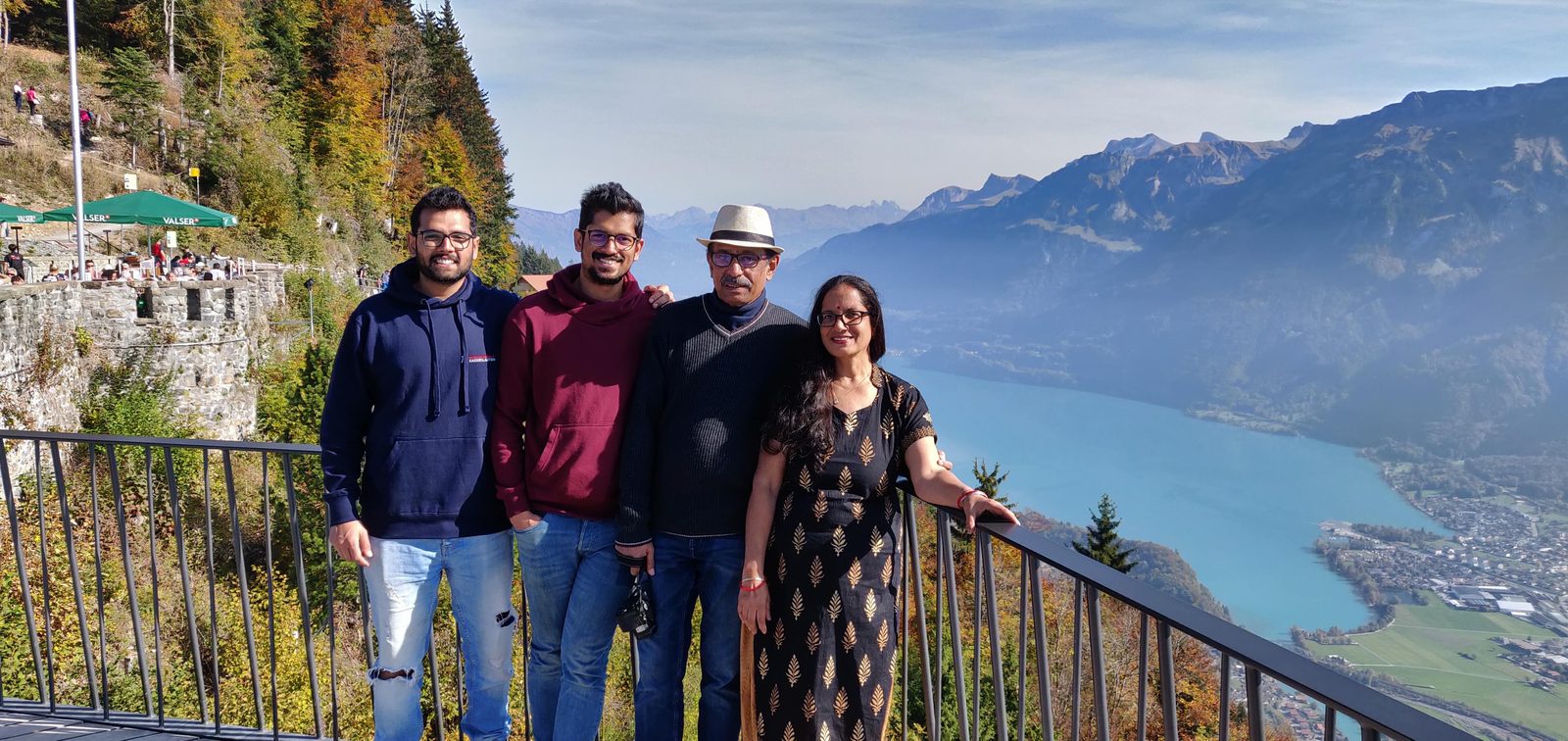 The founder's family at a railing above a turquoise lake: the founder on the left in a navy hoodie, his brother, his father in a hat, and his mother — the one who sends the morning messages — on the right in a black and gold kurta.
